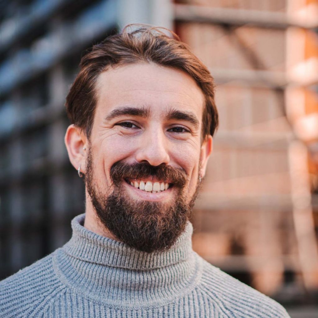 Porträt eines jungen Mannes mit braunem Haar und Vollbart, der freundlich lächelnd in die Kamera blickt. Er trägt einen grauen Rollkragenpullover und hat ein Ohrpiercing.