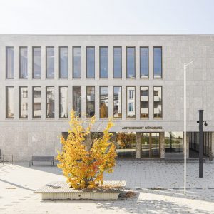 Modernes Gebäude des Amtsgerichts Günzburg mit heller Steinfassade und großen Fenstern, davor ein gepflasterter Platz mit einem Baum in herbstlicher gelber Laubfärbung. Der Eingangsbereich ist mit Glasfronten gestaltet, darüber der Schriftzug 'Amtsgericht Günzburg'.