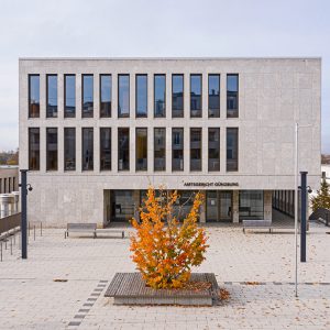 Das Amtsgericht Günzburg, ein modernes Gebäude mit einer Fassade aus hellem Stein und vertikalen Fensterreihen, dominiert einen gepflasterten Platz. Im Vordergrund steht ein Baum mit leuchtend orangefarbenem Herbstlaub.