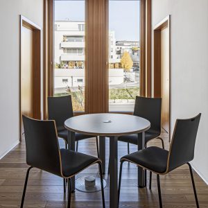 Ein minimalistischer Besprechungsraum mit vier schwarzen Stühlen um einen runden Tisch, platziert auf einem eleganten Holzboden. Im Hintergrund befinden sich hohe Holzrahmenfenster mit perforierter Verkleidung.