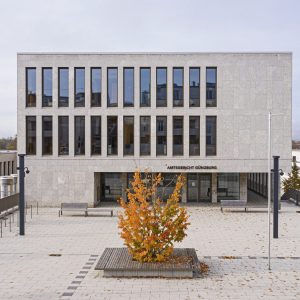 Moderne Fassade des Amtsgerichts Günzburg, einem kubischen Gebäude aus hellem Naturstein mit hohen, rechteckigen Fenstern in gleichmäßiger Anordnung. Der Eingangsbereich trägt den Schriftzug „AMTSGERICHT GÜNZBURG“. Im Vordergrund ein gepflasterter Vorplatz mit Bänken, einem Geländer und einem Baum in kräftigem Herbstlaub, der von einem rechteckigen Holzrahmen eingefasst ist.