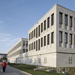Modernes Verwaltungsgebäude mit heller Steinverkleidung und rechteckigen, dunkel gerahmten Fenstern, das sich entlang eines gepflasterten Gehwegs erstreckt. Im Vordergrund eine Person mit roter Jacke, die mit einem Hund spazieren geht, bei klarem Himmel und spätnachmittäglichem Sonnenlicht.
