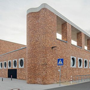 Ein markantes Backsteingebäude mit geschwungener Fassade, runden Fenstern und offenen rechteckigen Durchbrüchen im oberen Bereich. Im Vordergrund befinden sich ein Zebrastreifen, Fahrradständer und ein gepflasterter Vorplatz.