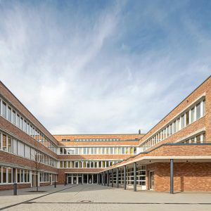 Ein moderner Schulhof mit einem U-förmigen Backsteingebäude, das große Fensterreihen und klare Linien aufweist. Der gepflasterte Vorplatz wird von einer überdachten Passage mit Stützsäulen ergänzt.
