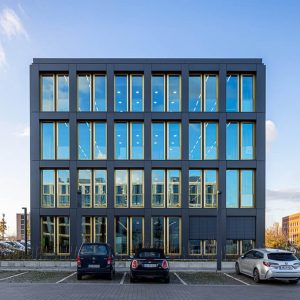 Moderne Bürogebäudefront mit vier Stockwerken, symmetrischer Glasfassade und dunklen Metallrahmen, die den blauen Himmel und umliegende Strukturen reflektieren. Drei Autos sind auf dem Parkplatz davor geparkt.
