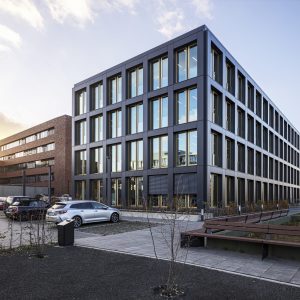 Ein modernes Bürogebäude mit dunkler Fassade und großen Fenstern mit goldenen Rahmen, aufgenommen bei Sonnenuntergang, wodurch ein warmes Licht die Umgebung beleuchtet. Im Vordergrund befinden sich ein Parkplatz mit mehreren Autos, eine Bank auf einem gepflasterten Weg und spärlich bepflanzte Grünflächen.