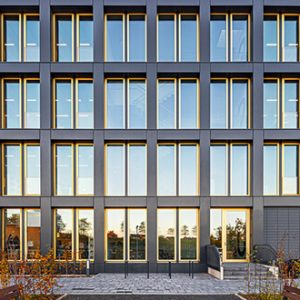 Frontansicht eines modernen Bürogebäudes mit symmetrischer Fensteranordnung, rechteckigen Glasflächen und dunklen Rahmen. Die Fassade reflektiert die Umgebung und das weiche Abendlicht.