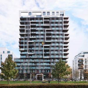 Ein modernes, mehrstöckiges Wohngebäude mit einer markanten Fassade aus Glas und geometrischen Balkonstrukturen, die ein spielerisches Muster aus Rechtecken bilden. Im Vordergrund befindet sich eine grüne Rasenfläche mit Bäumen.