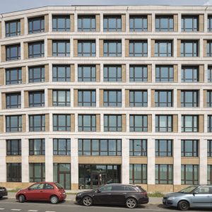 Ein symmetrisches, modernes Bürogebäude mit einer klaren Fassade aus weißen Steinrahmen, großen Glasfenstern und beigen Akzenten. Vor dem Gebäude stehen geparkte Autos entlang einer asphaltierten Straße.