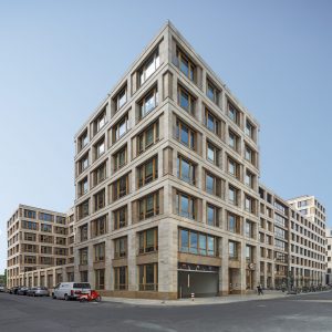Modernes Bürogebäude aus beigefarbenem Naturstein und großen Holzrahmenfenstern, das klare Linien und geometrisches Design vereint. Im Erdgeschoss befinden sich Eingänge und eine Tiefgarageneinfahrt, während Autos und Fahrräder entlang der Straße geparkt sind.