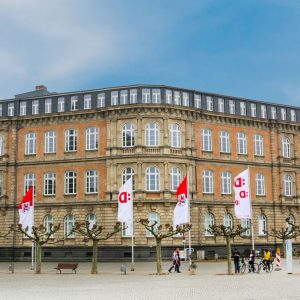 Historisches Gebäude mit roter Klinkerfassade und großen weißen Fenstern, verziert mit klassischen Steinelementen. Davor stehen kahle, symmetrisch angeordnete Bäume und mehrere Fahnen mit roten und weißen Logos.