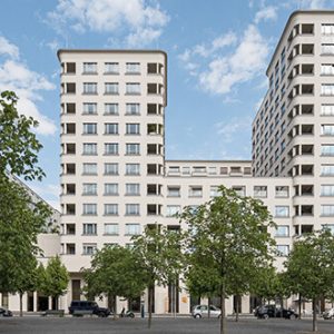 Frontalansicht eines modernen Gebäudekomplexes mit abgerundeten Ecken und heller Putzfassade, symmetrisch angeordneten Fenstern und klaren Linien. Im Vordergrund befinden sich eine Baumreihe, parkende Autos und ein breiter Gehweg.