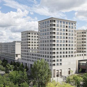 Panorama eines modernen Hotelkomplexes mit heller Fassade und symmetrischen Fenstern, umgeben von Bäumen, Parkanlagen und angrenzenden Stadtgebäuden.