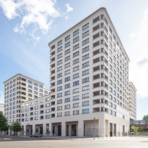 Moderne, weiß verputzte Hotelanlage mit abgerundeten Ecken und zahlreichen Fenstern, die eine klare und zeitgemäße Architektur präsentieren. Im Vordergrund befindet sich eine Straßenkreuzung mit Bäumen, Laternen und geparkten Autos, während die unteren Etagen Arkaden und Eingangspassagen zeigen.