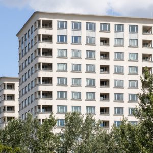 Nahaufnahme von zwei modernen Wohngebäuden mit weißen Fassaden und klar strukturierten Fenstern, die teilweise verdeckt sind von grünen Bäumen. Die Gebäude haben abgerundete Ecken und kleine Balkone an den Seiten.