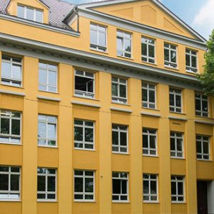 Gelbes mehrstöckiges Gebäude mit klassizistischen Elementen, symmetrischer Fassade und weißen Fensterrahmen umgeben von grünen Bäumen. Giebel und Dachfenster verleihen dem Bau ein charakteristisches Erscheinungsbild.