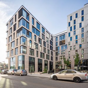 Ein modernes Gebäude mit heller Steinfassade, großen Glasfenstern und einer klaren, geometrischen Architektur, das sich über mehrere Etagen erstreckt. Im Vordergrund parken Autos entlang der Straße, während Bäume und blauer Himmel eine freundliche Atmosphäre schaffen.