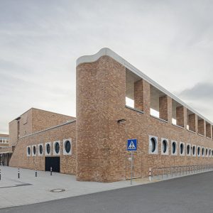 Ein langes Gebäude mit einer großen Backsteinfassade und runden Fenstern. Davor befinden sich ein Verkehrsschild für einen Fußgängerüberweg und zahlreiche Fahrradständer.