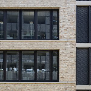 Nahaufnahme einer modernen Ziegelfassade mit großen, schwarzen Fenstern, die Einblicke in ein Bürointerieur geben. Rechts angrenzend sind vertikale, schwarze Metallpaneele mit perforiertem Muster zu sehen.