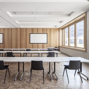 Moderner Bürobereich mit einem hohen schwarzen Tisch und passenden schwarzen Barhockern vor großen Holzrahmenfenstern, die den Blick auf ein benachbartes Gebäude freigeben. Im Hintergrund befinden sich Pflanzen in grauen Pflanzgefäßen und eine Sitzecke mit minimalistischen Möbeln.