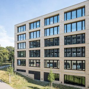 Moderne Gebäudefassade aus hellen Ziegelsteinen mit großen, symmetrisch angeordneten Fenstern und klarer, geradliniger Architektur. Vor dem Gebäude verläuft ein begrünter Weg mit jungen Bäumen und gepflegter Vegetation.
